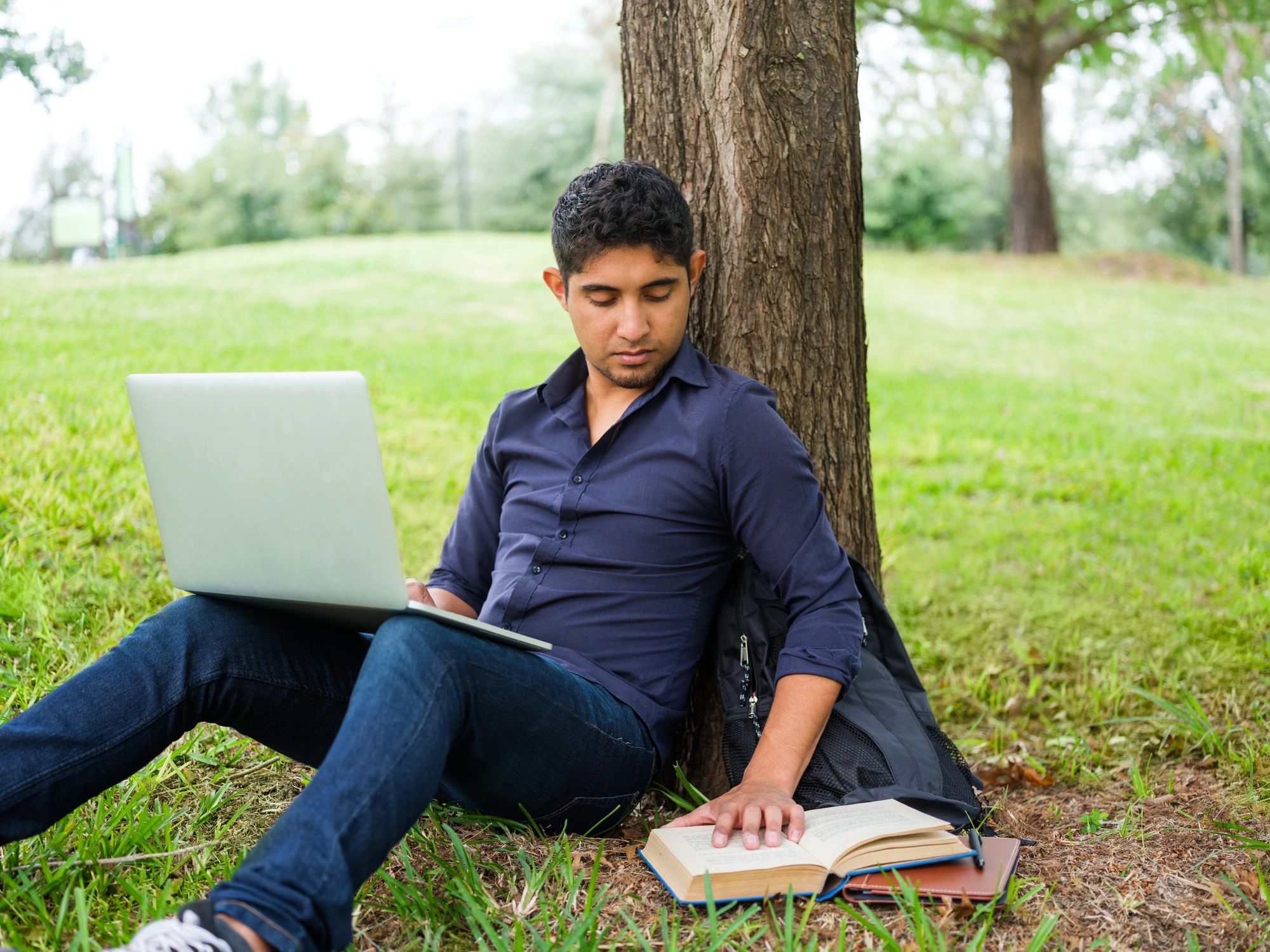 Man reading a book while using a laptop outdoors